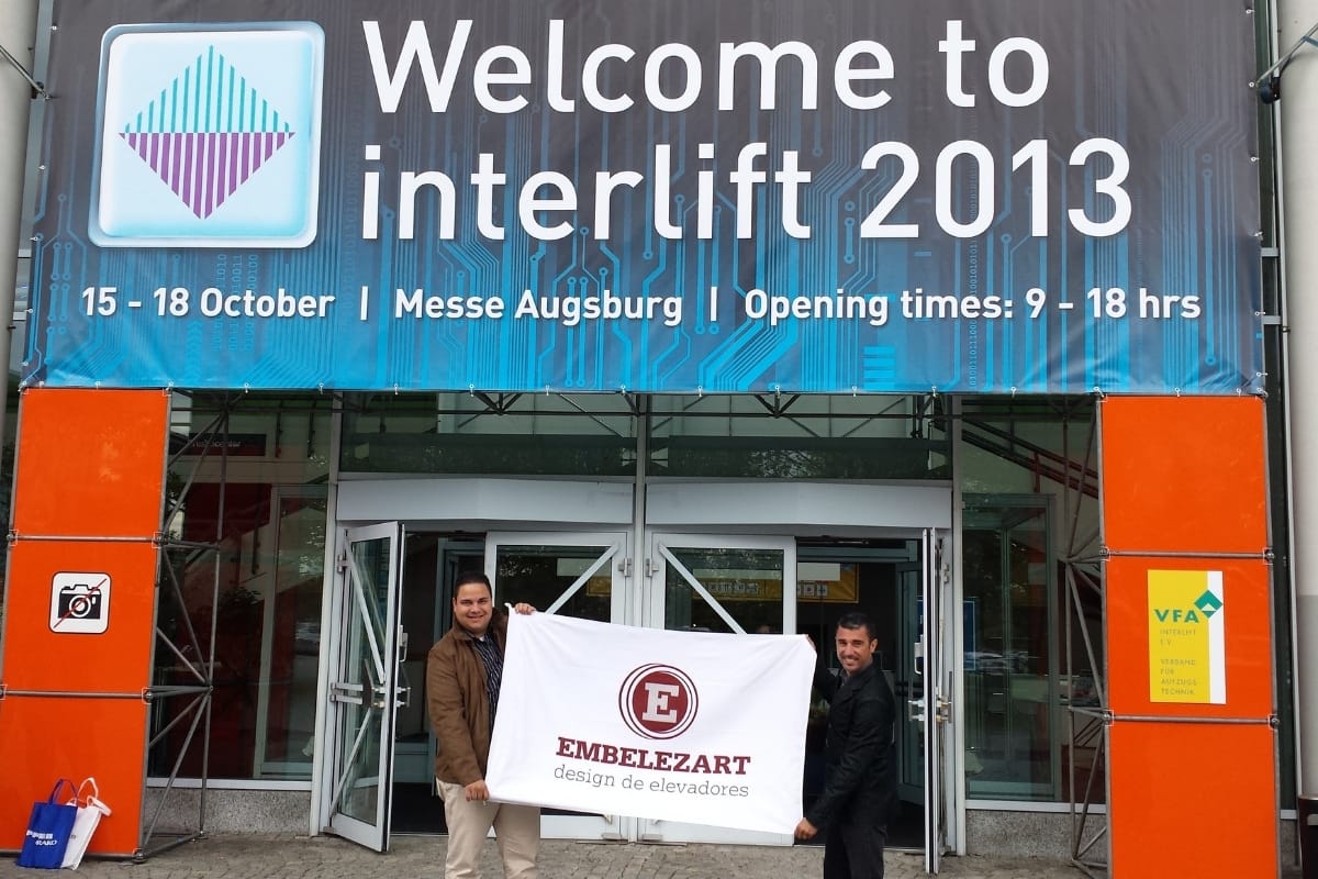 Equipe da Embelezart Elevadores com a bandeira da empresa na entrada da feira Interlift 2013, em Messe Augsburg, Alemanha
